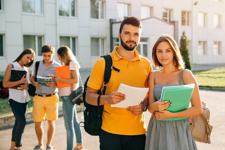 Two happy students with backpacks and books in their hands smiling at camera while standing on background of university and friends